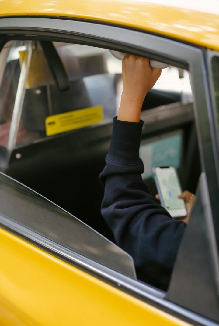 Unrecognizable passenger sitting on backseat and browsing smartphone while holding handle inside of yellow cab with lowered window during ride